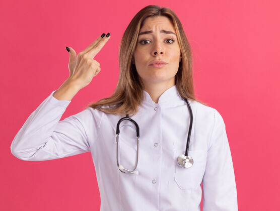 年轻人年轻的女医生穿着医用长袍 听诊器显示的手势 隔离在粉红色的墙上穿着感情