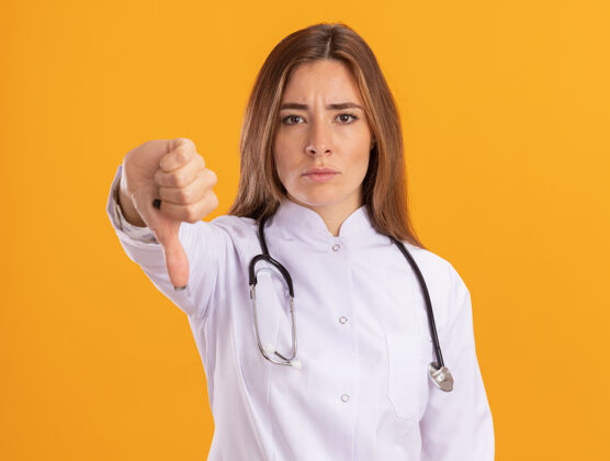 表情严格的年轻女医生穿着医用长袍 听诊器显示拇指向下隔离在黄色的墙上听诊器衣服下