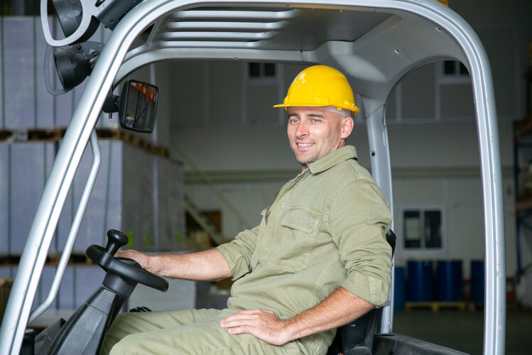 人戴着安全帽的快乐男仓库工人的画像在仓库里驾驶叉车 握着方向盘 微笑着 望着别处生产设备叉子