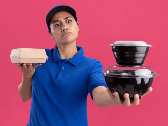 容器严格的年轻送货女孩穿着制服 戴着帽子 把食物容器隔离在粉红色的墙上帽子穿食品