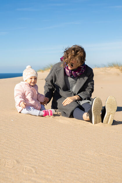 休闲快乐的妈妈和可爱的小宝宝穿着暖和的衣服 在海上度过闲暇时光 一起坐在沙滩上家庭父母欢呼