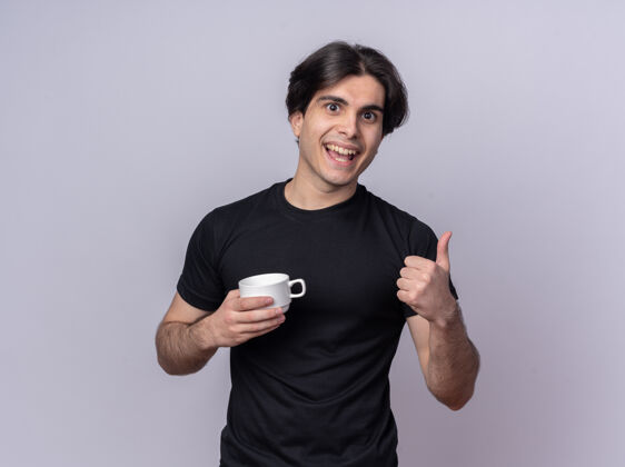 帅气面带微笑的年轻帅哥穿着黑色t恤 手里拿着一杯咖啡 在白色的墙上孤立地竖起大拇指咖啡穿拇指