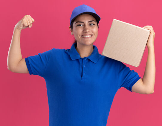 帽子微笑的年轻送货女孩穿着制服 肩上扛着帽子盒 在粉红色的墙上显示出强烈的姿态抱着年轻肩膀