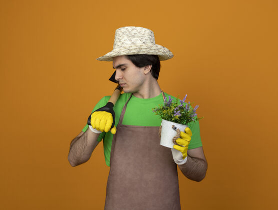 持有身穿制服的年轻男园丁戴着园艺帽戴着手套闭着眼睛拿着花盆里的花把铁锹放在肩膀上隔离在橘子上男橘子手套