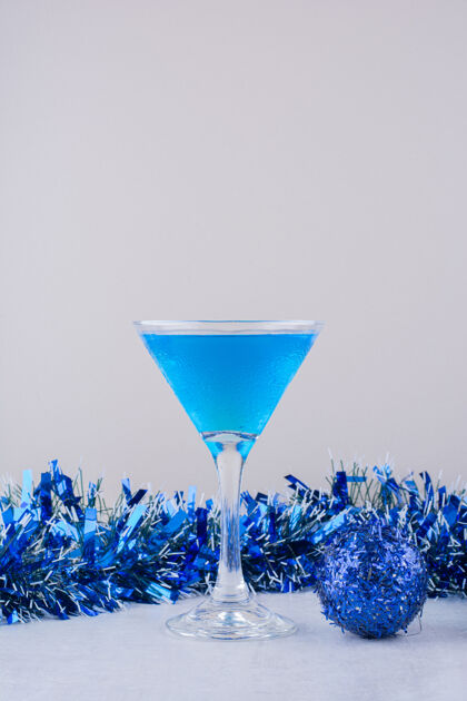 鸡尾酒一杯蓝色鸡尾酒 旁边是白色表面上的蓝色圣诞装饰品舞会花圈圣诞