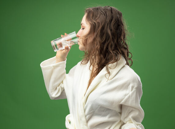 年轻站在侧视图上的年轻生病的女孩穿着白色长袍 喝着绿色隔离的水绿色水疾病