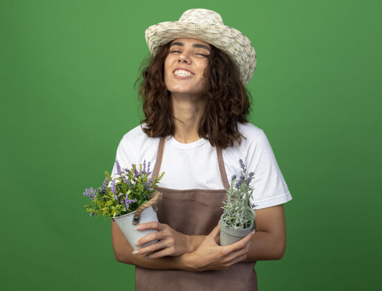 举行微笑着的年轻女园丁 穿着制服 戴着园艺帽 手拿鲜花 在绿色的花盆里交叉女性花盆穿