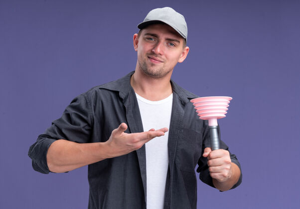 点高兴的年轻帅气的清洁工穿着t恤 戴着帽子拿着安斯点着隔离在紫色墙上的柱塞年轻柱塞请
