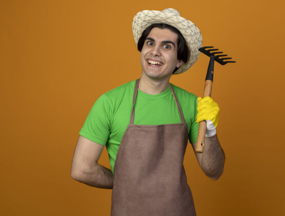 耙子快乐的年轻男园丁穿着制服 戴着园艺帽 戴着手套 手里拿着一把橙色的耙子男性手套持有