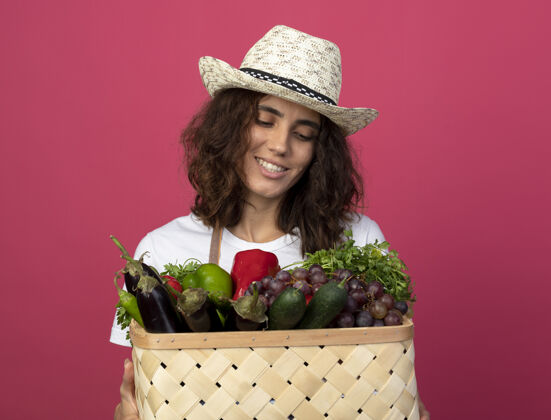 帽子年轻的女园丁面带微笑 穿着制服 戴着园艺帽 拿着一只粉红色的菜篮子望着园艺穿粉红色