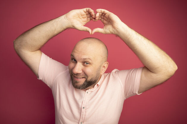 男人微笑的中年秃头男子穿着粉色t恤举着心形孤立的粉色秃顶穿手势