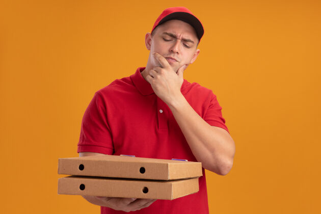 下巴年轻的送货员穿着制服 戴着帽子 看着披萨盒 手放在橘色墙上的下巴上盒子披萨手
