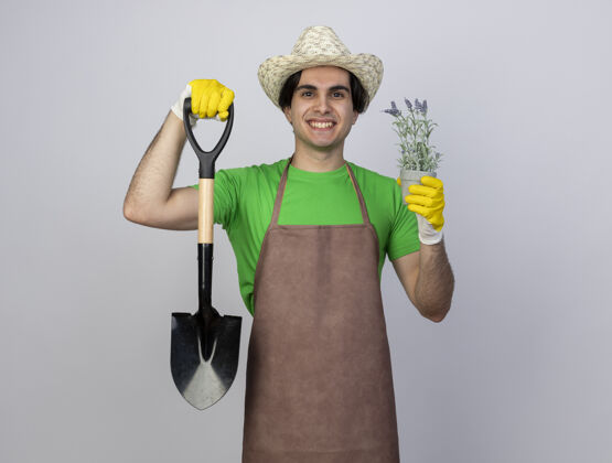 帽子微笑着的年轻男园丁 穿着制服 戴着园艺帽和手套 手里拿着花盆里的花的铁锹花制服抱