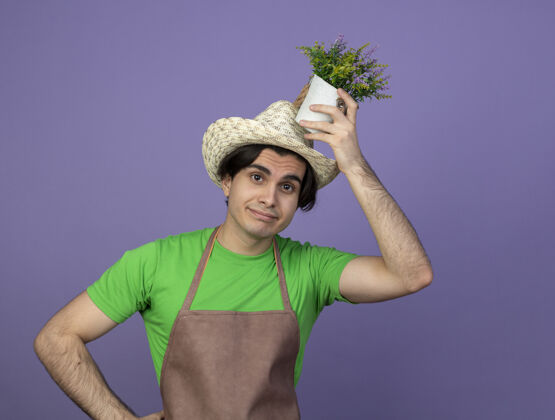 紫色高兴的年轻男园丁穿着制服 戴着园艺帽 头上捧着花盆里的花男人穿花盆