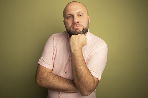 粉色自信的中年秃头男人穿着粉色t恤把手放在下巴下孤立的橄榄绿上橄榄色绿色男士