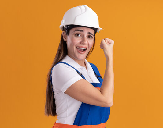 年轻身着制服的快乐的年轻建设者女子在橙色的墙上展示着坚强的姿态姿态建设者表演