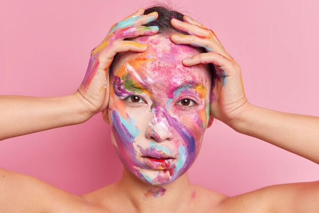 化妆严肃的深色头发的亚洲女人的头像手放在头上直接看着涂有颜料的相机在粉色背景下赤裸的肩膀摆出姿势女性彩色严肃