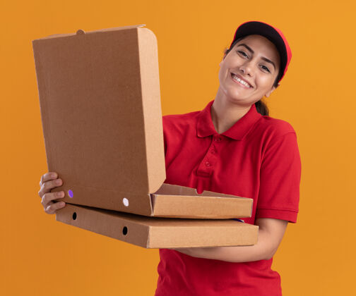 披萨微笑的年轻送货女孩穿着制服 戴着帽子 拿着披萨盒 打开披萨盒 隔离在橙色的墙上送货微笑年轻