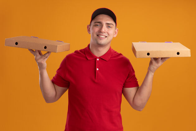 微笑微笑着的年轻送货员穿着制服 戴着帽子 拿着隔离在橙色墙上的比萨饼盒帽子年轻人拿着