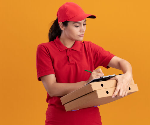 穿着自信的年轻送货女孩穿着制服 戴着帽子 在剪贴板上写着什么 就在隔离在橙色墙上的比萨饼盒上女孩披萨盒子