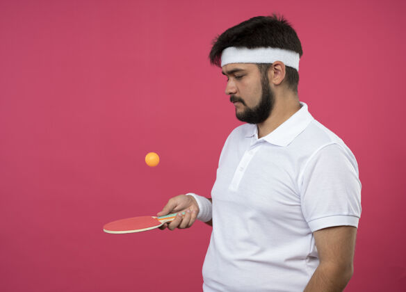 穿戴着头带和腕带 拿着乒乓球拍 把球孤立在粉红色墙上的年轻运动型男子腕带年轻男子