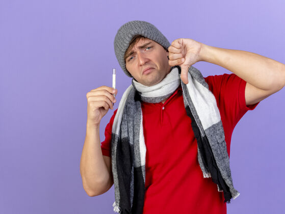 姿势年轻英俊的金发病男子戴着冬天的帽子和围巾拿着温度计看前面显示拇指向下孤立在紫色墙上复制空间衣服人拇指