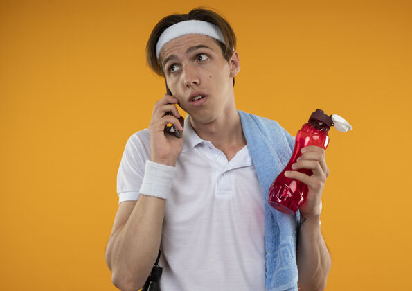 电话令人印象深刻的年轻的运动型家伙看着一边戴着头带和手带与毛巾肩上讲电话隔离在橙色的墙壁头巾架子年轻人