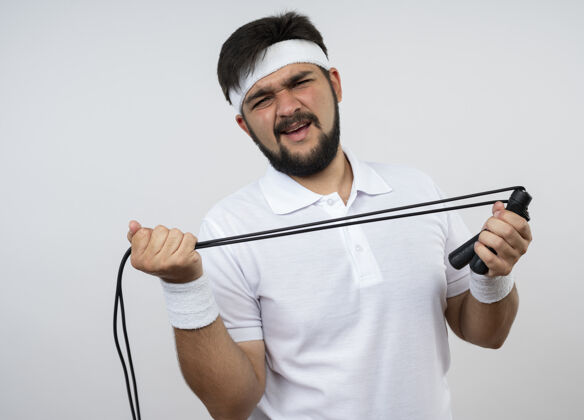 伸展戴着头带和腕带的未被释放的运动型年轻人在白色的墙上拉着一根跳绳运动头带不愉快