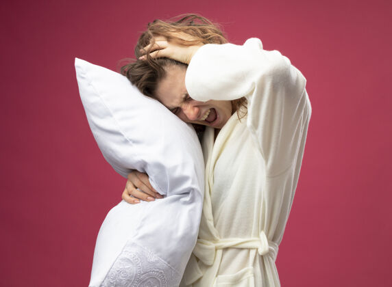抓住疼痛的年轻生病的女孩穿着白色长袍抱住枕头抓着头孤立在粉红色长袍年轻枕头