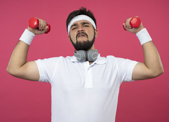 粉红自信的年轻运动型男子戴着头带和腕带 戴着耳机 用粉色的哑铃练习锻炼年轻训练