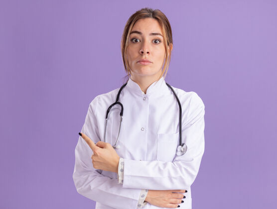 年轻惊讶的年轻女医生穿着带听诊器的医用长袍点在一边 隔离在紫色的墙上 留有复印空间医疗听诊器长袍