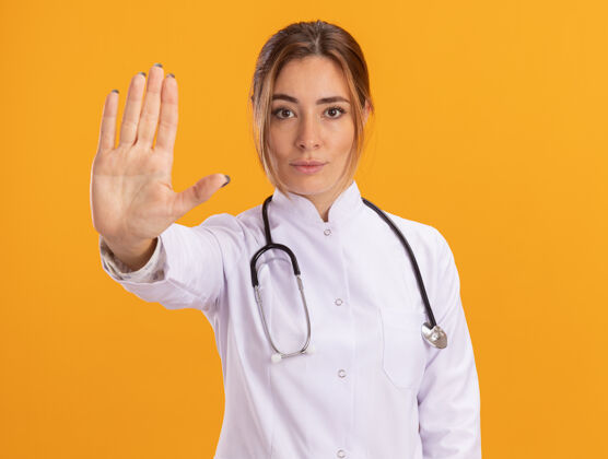 女性自信的年轻女医生穿着医用长袍 听诊器显示停止手势 隔离在黄色墙上停止医疗穿着