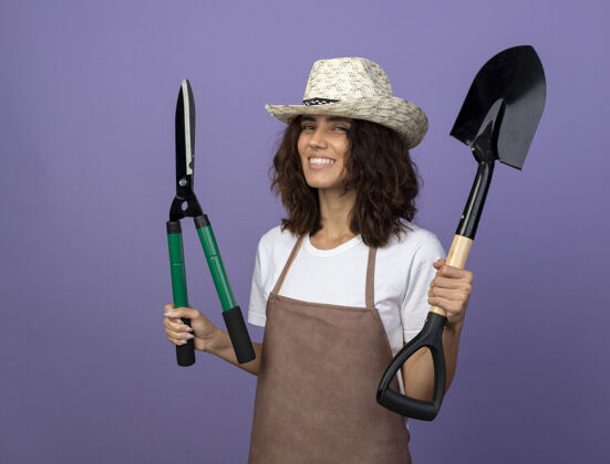 人身着制服的年轻女园丁微笑着戴着园艺帽 用剪刀举起铁锹穿女人帽子