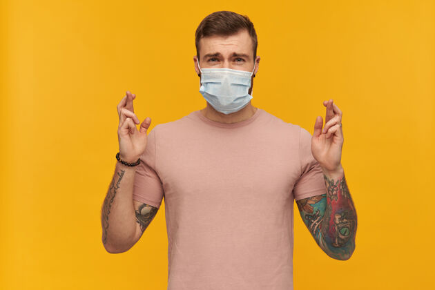 药困惑的年轻人穿着粉色T恤 脸上戴着防病毒面具 留着胡须和纹身 手指交叉着 在黄色的墙上许愿安全纹身T恤