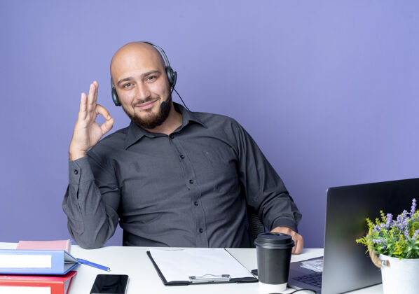 请戴上高兴的年轻秃头呼叫中心的男子戴着耳机坐在办公桌旁与工作工具做ok标志隔离紫色背景耳机年轻秃头