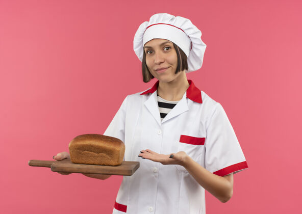请身着厨师制服的年轻女厨师手拿着一块粉红色背景的面包 指着砧板 高兴极了制服板年轻
