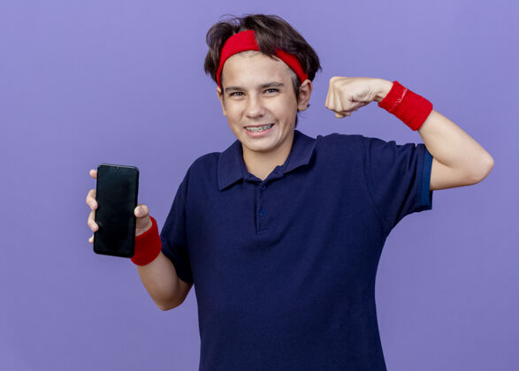 表情快乐的年轻帅气的运动男孩戴着头带和护腕 戴着牙套 手机做着强烈的手势 看着紫色墙壁上孤立的前方姿势头带运动