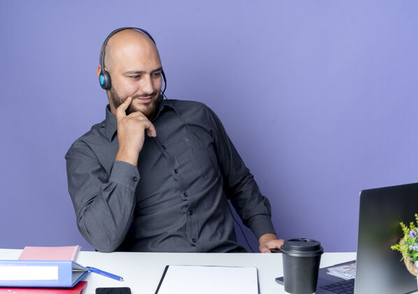 年轻年轻秃顶的呼叫中心男子戴着耳机坐在办公桌旁 工作工具被隔离在紫色的背景上坐着穿工具