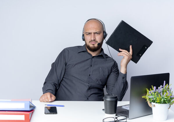 磨损戴着耳机的秃头呼叫中心年轻人坐在办公桌旁 手里拿着工作工具 把剪贴板隔离在白色背景上电话工具秃头