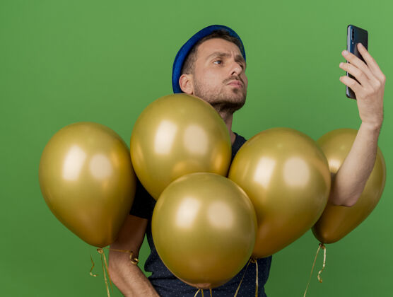 帅气自信的帅哥戴着蓝色的派对帽站着 手里拿着氦气球 看着隔离在绿色墙上的复制空间表情衣服感觉