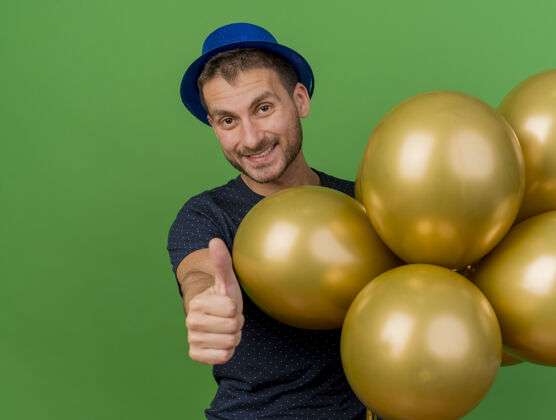 表情微笑的帅哥戴着蓝色的派对帽 手持氦气球 竖起大拇指孤立地站在绿色墙壁上 留着复制空间人人站立