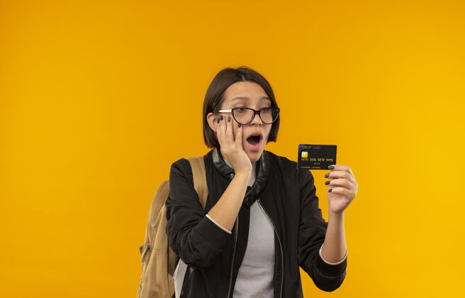 脸印象深刻的年轻女学生戴着眼镜 背着包拿着信用卡 手放在脸上孤立的橙色包持有眼镜