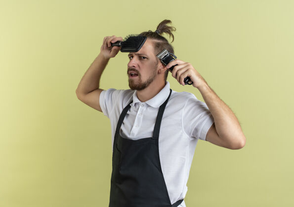年轻年轻帅气的理发师拿着梳子和理发器 靠近头发 橄榄绿背景 留有复印空间近头发剪子