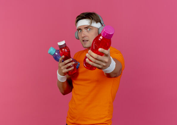 穿印象深刻的年轻英俊的运动男子戴着头带和腕带和耳机举行和伸出水瓶孤立的粉红色印象粉色运动
