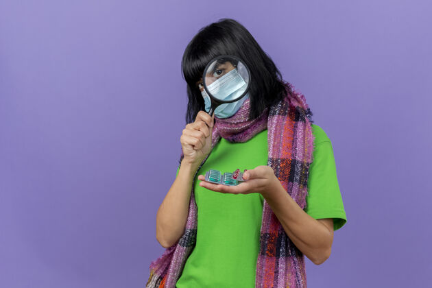 玻璃戴着口罩和围巾的年轻病妇拿着一包包胶囊透过放大镜看前面 隔离在紫色的墙上 留着复印空间胶囊人包