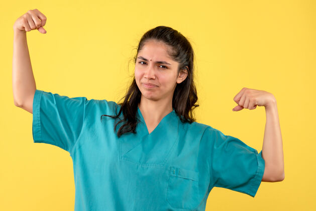 微笑前视图穿着制服的女医生展示她的手臂肌肉站立年轻人黄色