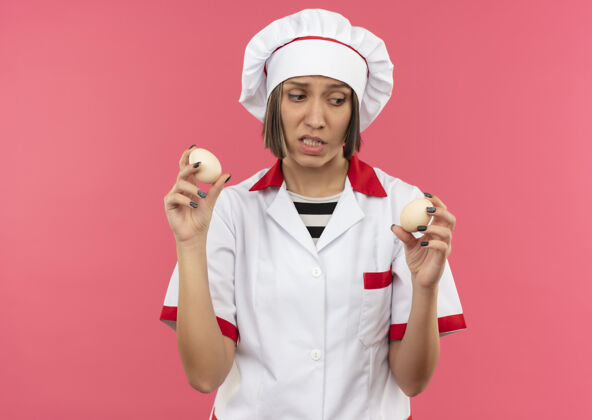 烹饪穿着厨师制服的年轻女厨师手拿鸡蛋 看着粉红色背景上孤立的鸡蛋制服年轻女