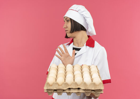 年轻穿着厨师制服的年轻女厨师拿着一盒鸡蛋 一边看一边做手势 粉色背景上没有单独的复制空间烹饪鸡蛋不愉快