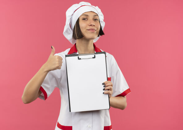 拿着高兴的年轻女厨师在厨师制服举行剪贴板和显示拇指上孤立的粉红色背景与复制空间拇指剪贴板年轻