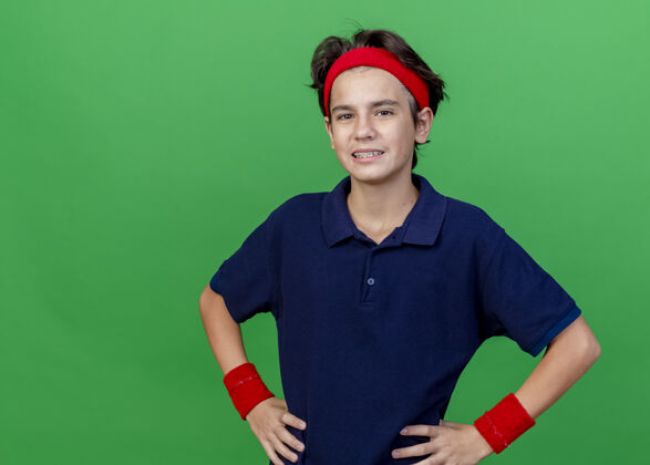 衣服微笑的年轻帅气的运动男孩戴着头带和护腕 戴着牙套 手放在腰上 看着前面 隔离在绿色墙壁上 留着复制空间人手帅气